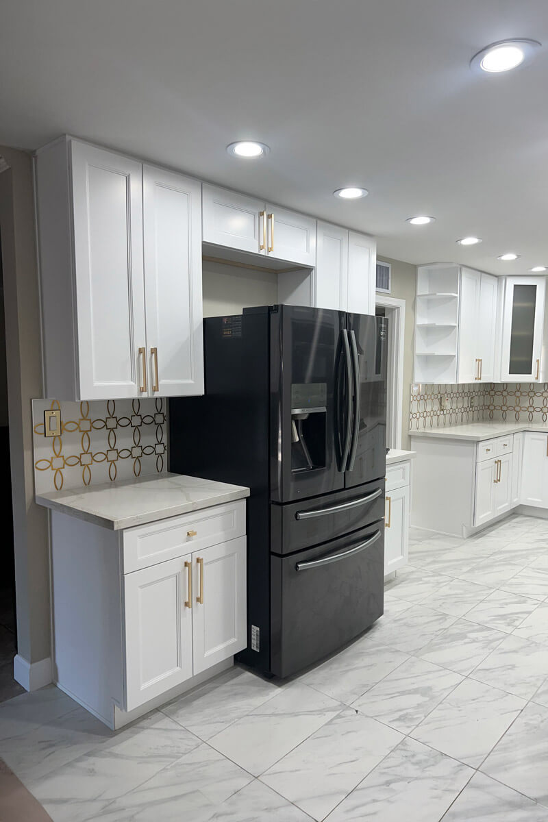 A bright white kitchen featuring marble-style floors, black appliances, and gold-accented hardware. Recessed lights highlight the clean lines and spacious layout.