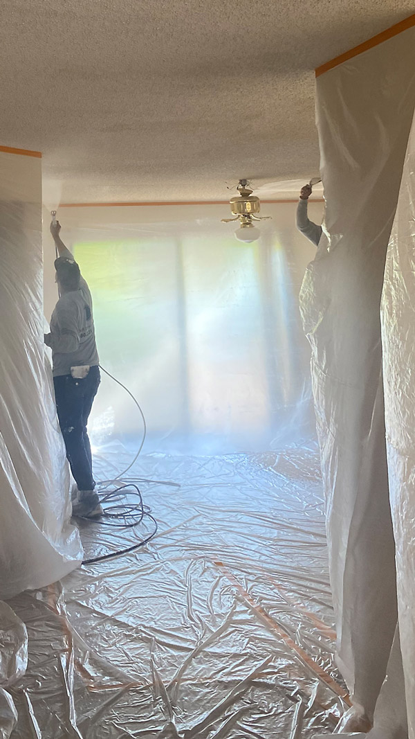 Workers prepare a room for ceiling work by covering the walls and floor with plastic sheeting.