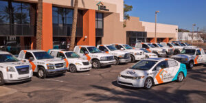 A fleet of white service vehicles branded with the “JD Lux Improvements” logo parked in front of a modern commercial building. The vehicles include trucks, vans, and a car, all featuring orange and teal graphics, representing a professional property improvement company.