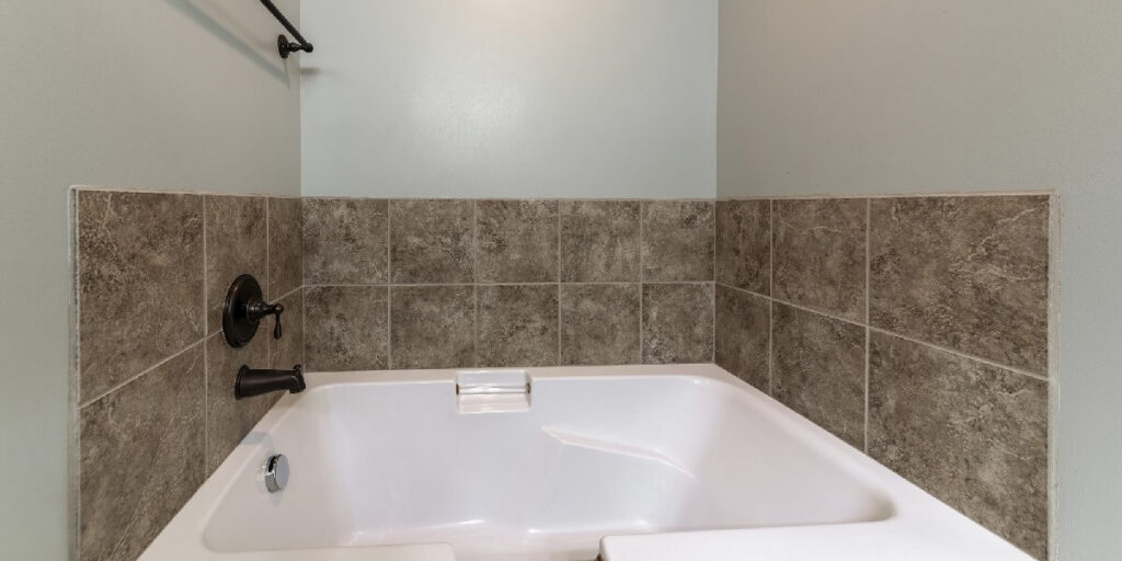 A clean white soaking tub set into stone-look gray tile surround with dark bronze fixtures in a residential bathroom.
