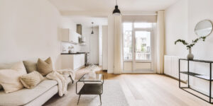 Bright minimalist living room with a beige sofa and open kitchen leading to a balcony door.